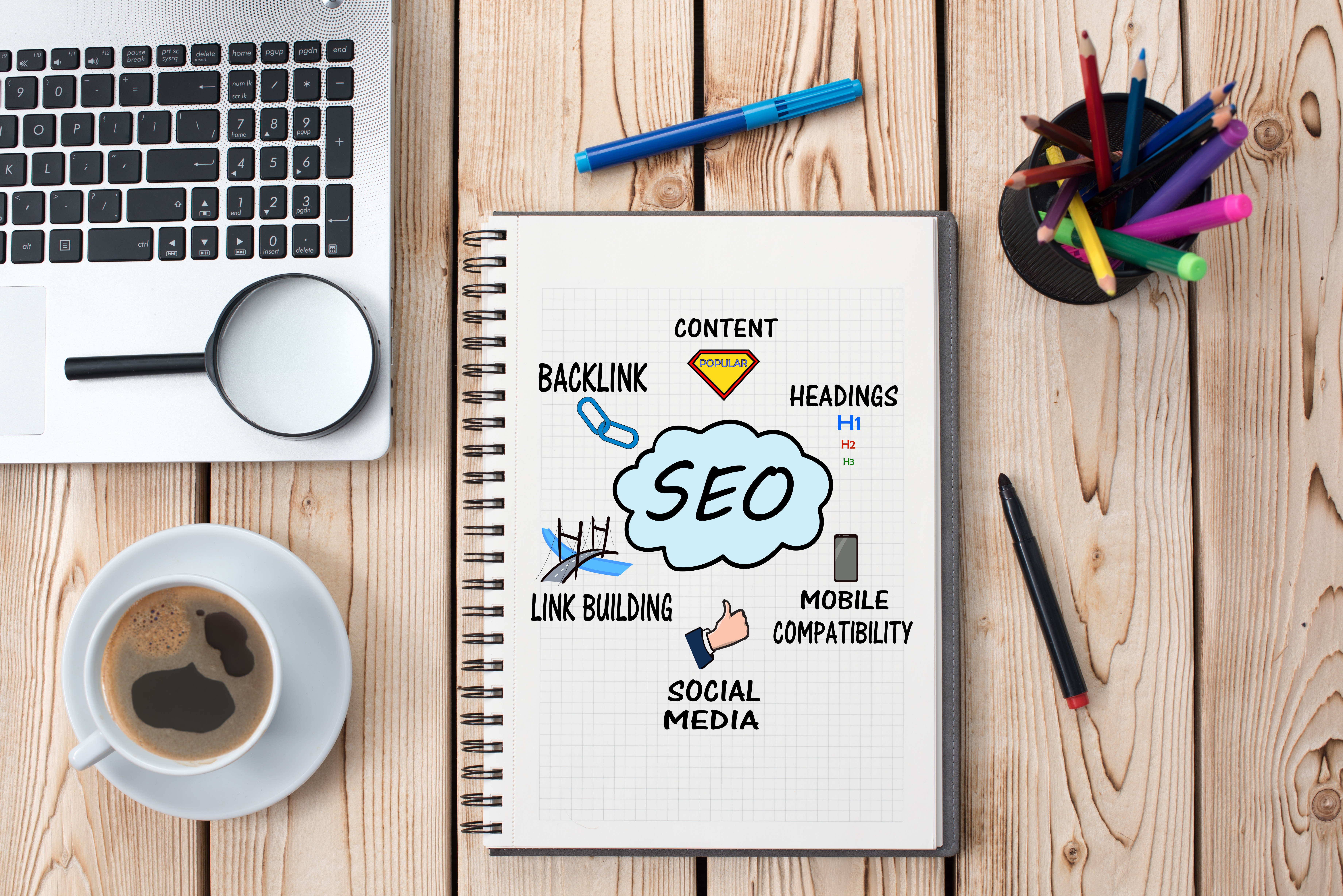 An open notebook with a written word “SEO” on its page is lying on a table next to a cup of coffee and open laptop.  Brainstorm solutions are drawn around the word “SEO” on a notebook’s page. 